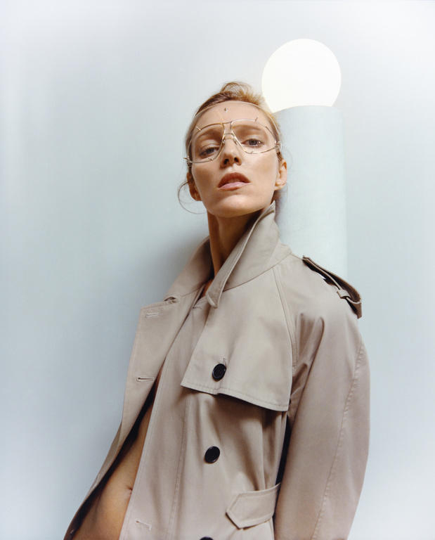 A woman in a trench coat and gold glasses leans against a column with a glowing orb in a fashion forward, indoor portrait.