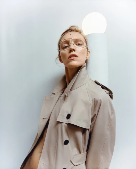 A woman in a trench coat and gold glasses leans against a column with a glowing orb in a fashion forward, indoor portrait.