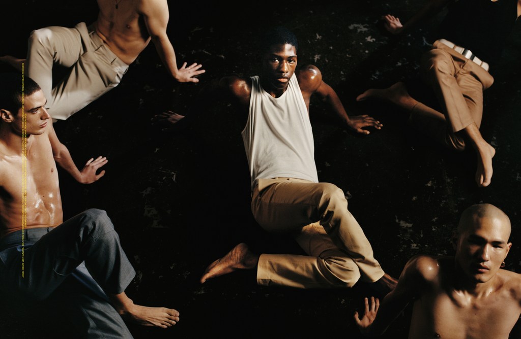 Group of shirtless men in trousers sit on a dark surface in a studio, lit with a soft light.