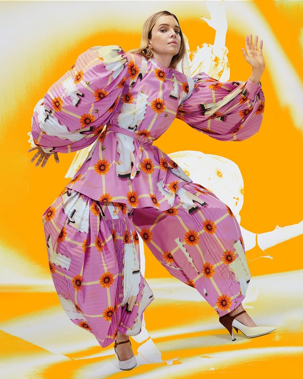 A woman poses in an eye-catching pink plaid pantsuit with sunflower and feather accents against an abstract orange background.