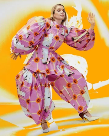 A woman poses in an eye-catching pink plaid pantsuit with sunflower and feather accents against an abstract orange background.