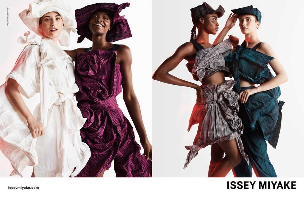 Two female models in draped fabric outfits and matching hats pose in a brightly lit studio against a seamless white background.
