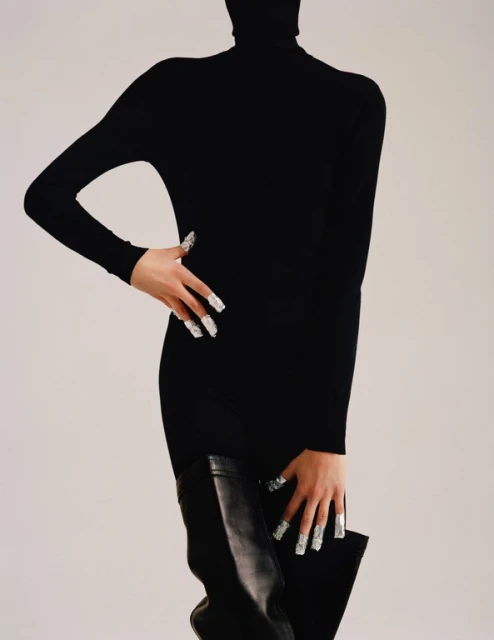 Fashionable woman poses indoors wearing a long-sleeved black turtleneck dress, leather thigh-high boots, and silver manicured nails. 