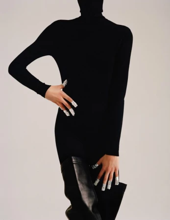 Fashionable woman poses indoors wearing a long-sleeved black turtleneck dress, leather thigh-high boots, and silver manicured nails. 