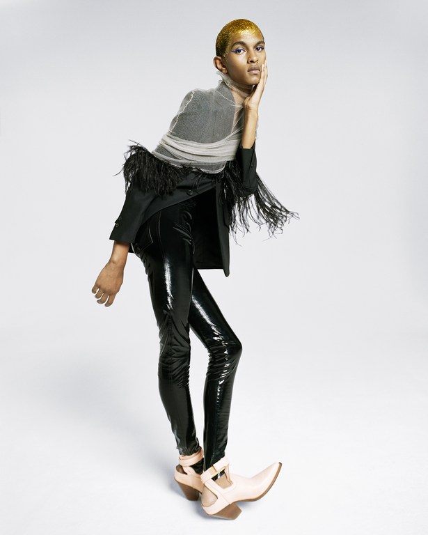 Model with gold glitter hair poses in black blazer with feather details, sheer top, shiny black leggings, and unique cutout heels in studio.