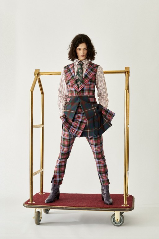 A woman in a multicolored plaid suit stands on a luggage cart against a white backdrop in a fashion editorial style photograph.