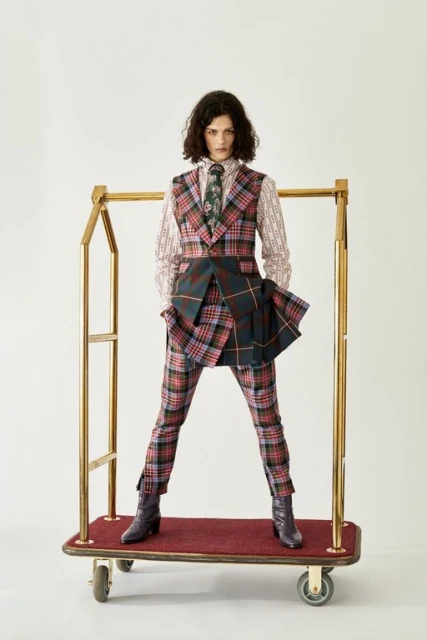 A woman in a multicolored plaid suit stands on a luggage cart against a white backdrop in a fashion editorial style photograph.