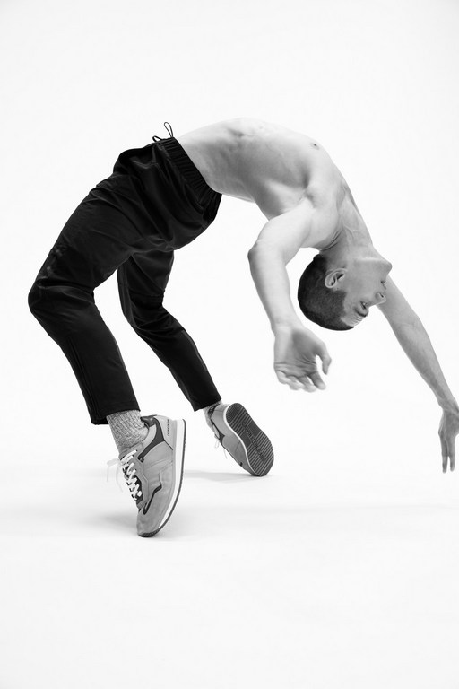 Monochrome image of a slender man in a backbend, wearing dark sweatpants and sneakers, on a bright white seamless background.