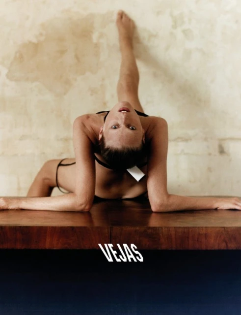 An adult female is reclining on a wooden table wearing black swimwear, while a tan wall provides a neutral backdrop in a portrait.