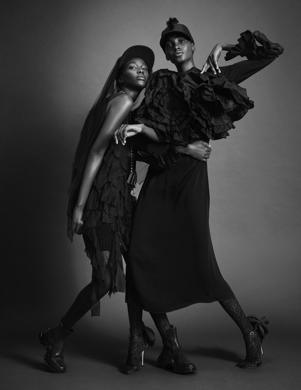 Two black women pose in black ruffled dresses and caps in a studio against a grey backdrop for a fashion shot, exuding gothic style.