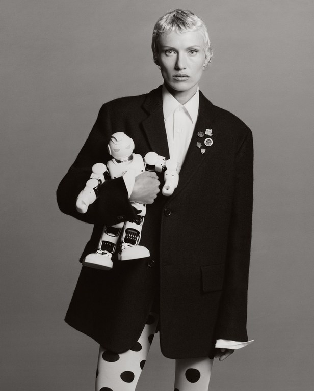 Monochrome portrait of a light-skinned adult female in a black blazer, holding a white robot toy against a solid grey backdrop.