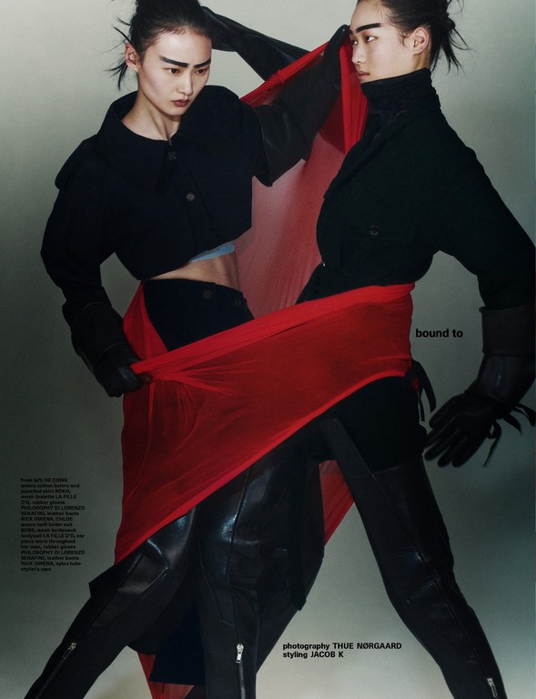 Two fashion models pose together in a photo studio wearing black jackets, gloves, and thigh-high boots, connected by a red fabric.