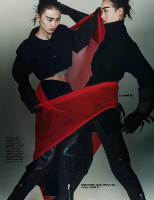 Two fashion models pose together in a photo studio wearing black jackets, gloves, and thigh-high boots, connected by a red fabric.