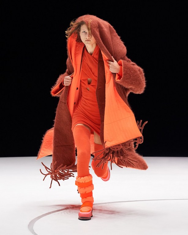 Runway fashion: a model presents an oversized brown fringe coat and bright orange dress with matching leg warmers and platform boots.