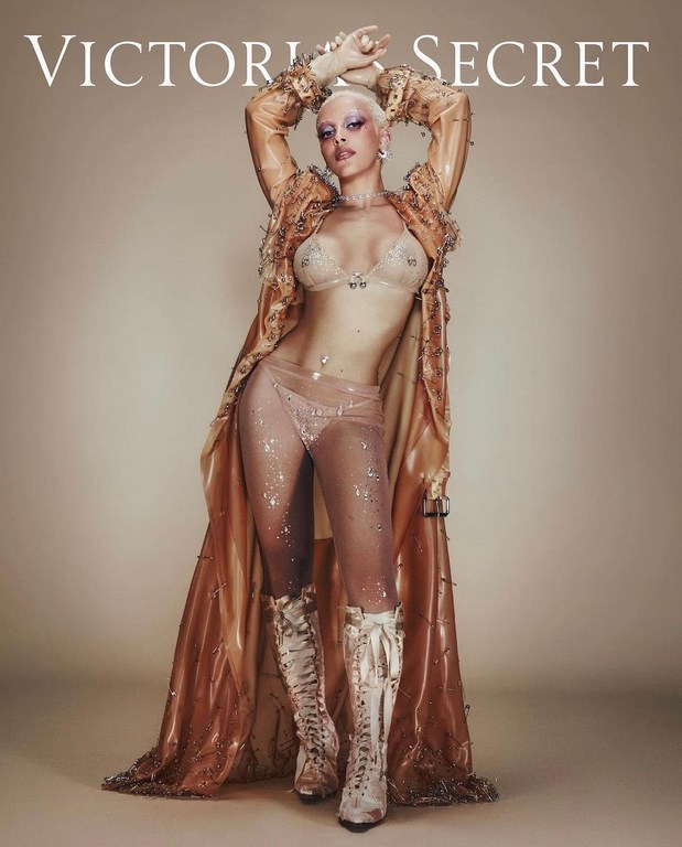 Woman with shaved head in transparent fashion posing indoors for Victoria's Secret, wearing lingerie, cape, and boots. Beauty and fashion.