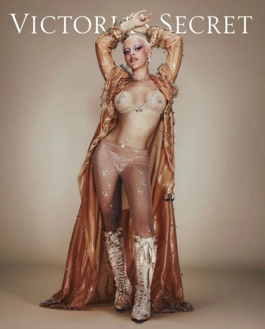 Woman with shaved head in transparent fashion posing indoors for Victoria's Secret, wearing lingerie, cape, and boots. Beauty and fashion.