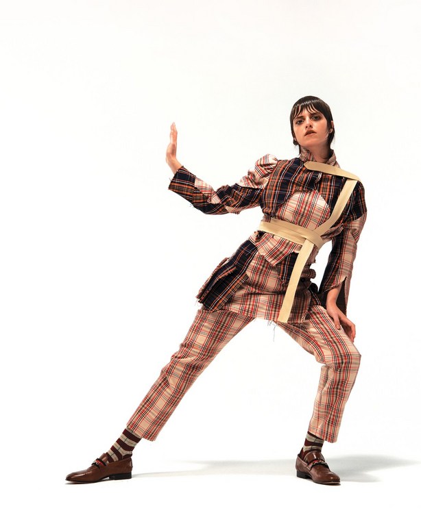 Model poses in a plaid suit with brown shoes and socks against a stark white backdrop in a fashion editorial-style photograph.