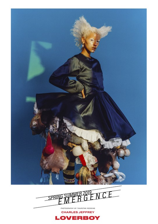 Fashion photo featuring a model in an avant-garde dark-blue dress adorned with plush objects. The backdrop is a solid light-blue.