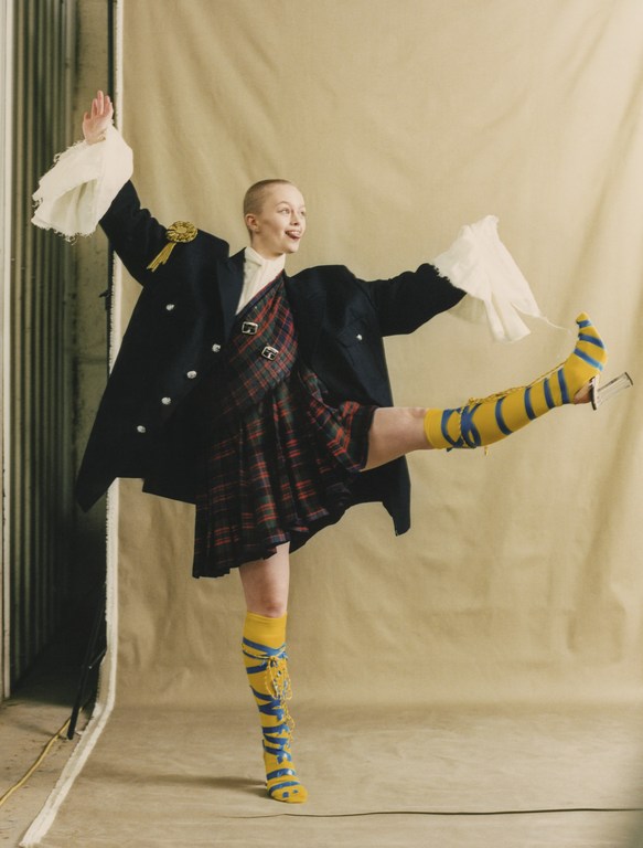 An adult woman in a black coat and red tartan kilt raises her leg up and sticks her tongue out in a studio environment.