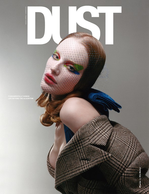 Model with red hair wearing a plaid blazer and mesh face veil against a neutral studio background