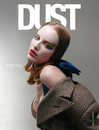 Model with red hair wearing a plaid blazer and mesh face veil against a neutral studio background