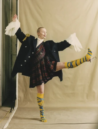 Dancer with a shaved head in a dark blazer and plaid dress extending a leg wearing yellow striped boots
