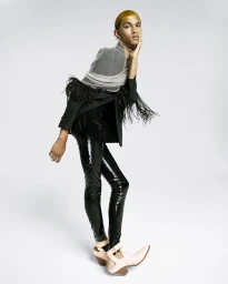 Model with gold glitter hair wearing black blazer, shiny pants and pink cutout boots against white studio background