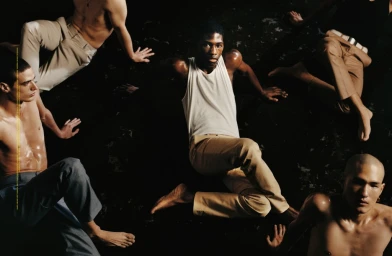Group of men posed dramatically on a dark surface, featuring a central figure in a white tank top and tan trousers.