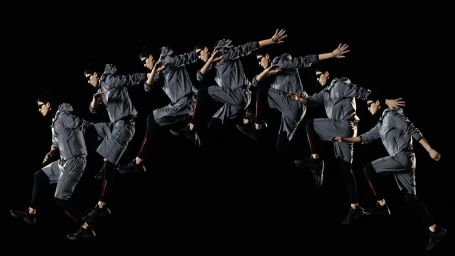 Multi-exposure action sequence of a male dancer in grey athletic wear and sunglasses leaping across a black background.