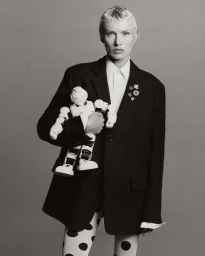 Monochrome portrait of a person with short blonde hair wearing an oversized black blazer holding a white toy robot