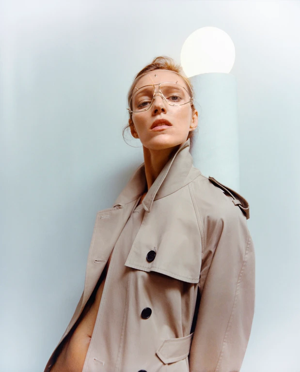 Portrait of a woman wearing wireframe glasses and a beige trench coat against a pale wall with a round light fixture.