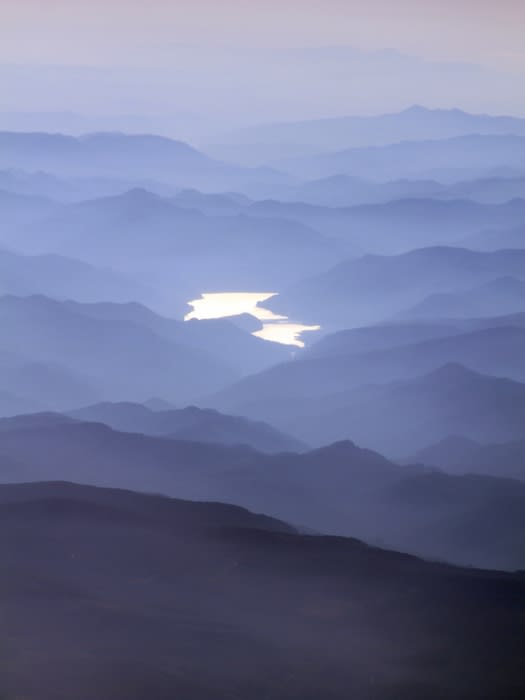 Aerial view of misty mountain ranges with a winding river reflecting light, creating a serene and layered landscape.