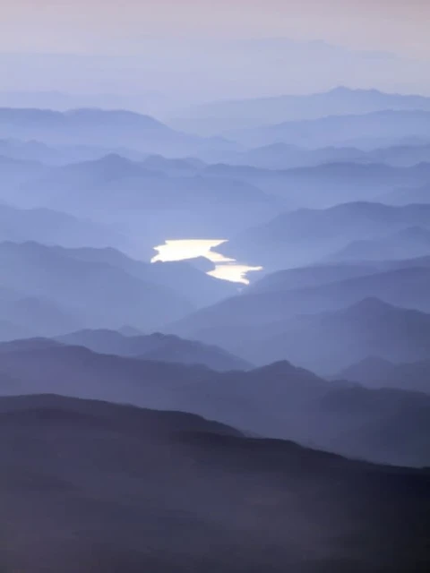 Aerial view of misty mountain ranges with a winding river reflecting light, creating a serene and layered landscape.