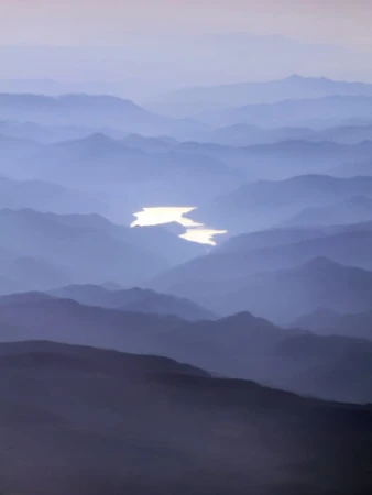 Aerial view of misty mountain ranges with a winding river reflecting light, creating a serene and layered landscape.