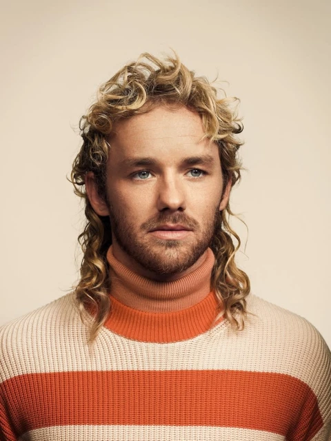 Portrait of a blond-haired man with a mullet wearing a striped orange and cream sweater against a plain background in a close-up studio shot.