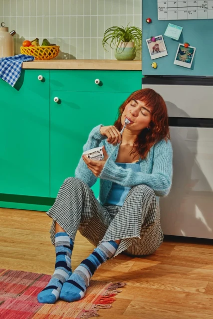 A woman sits on the floor in a kitchen and eats from a container with a spoon. She wears casual clothing and colorful socks.
