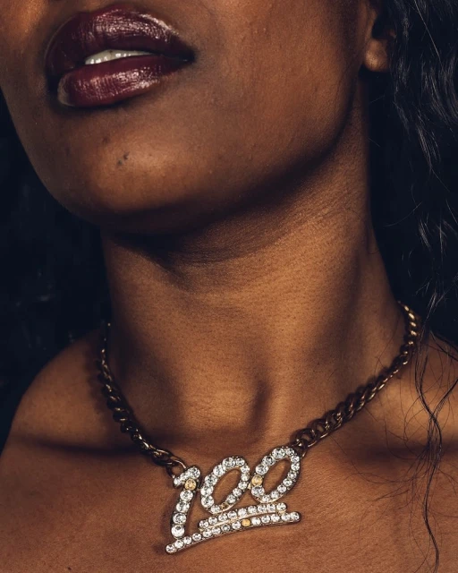 Close-up of a woman with dark skin and glossy dark lipstick wearing a gold chain with a large '100' pendant. Hair is dark and wavy.
