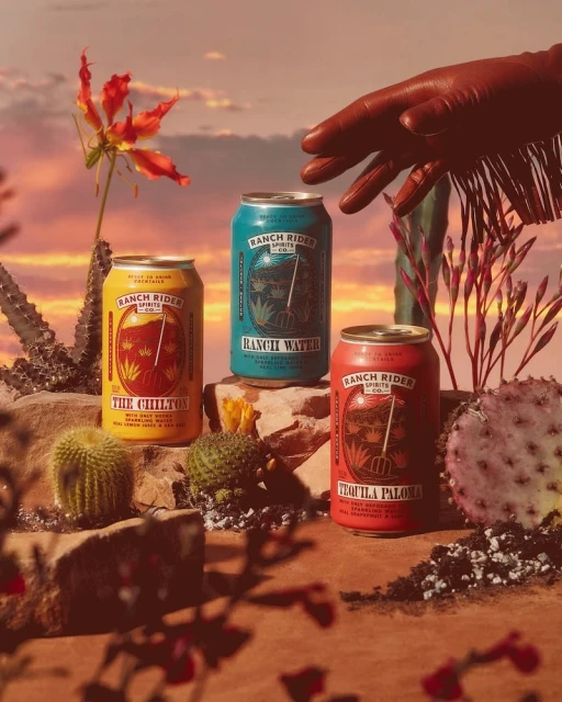 Three cans of Ranch Rider Spirits stand among desert flora under a sunset sky, with a gloved hand reaching towards one.