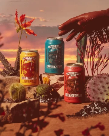 Three cans of Ranch Rider Spirits stand among desert flora under a sunset sky, with a gloved hand reaching towards one.