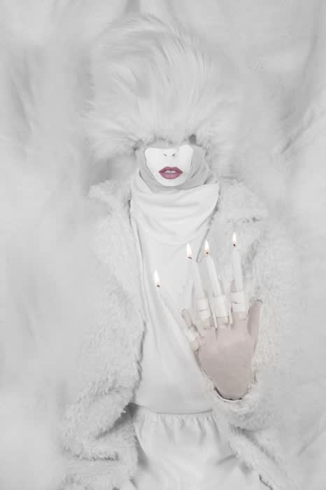 Abstract portrait of a person in white clothing with lit candles on their hand, set against a white background with theatrical fluff.