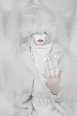 Abstract portrait of a person in white clothing with lit candles on their hand, set against a white background with theatrical fluff.
