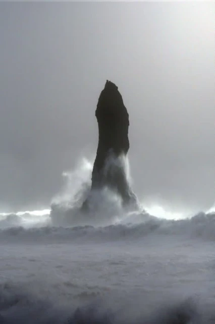 Monochromatic landscape featuring a tall, rocky sea stack rising from the ocean waves under an overcast sky, creating a dramatic scene.