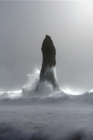Monochromatic landscape featuring a tall, rocky sea stack rising from the ocean waves under an overcast sky, creating a dramatic scene.