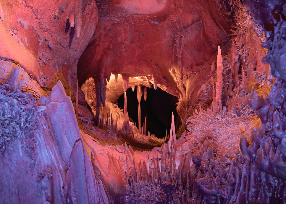 View of a cave with rock formations in hues of red and purple, illuminated by artificial light, creating a surreal landscape.