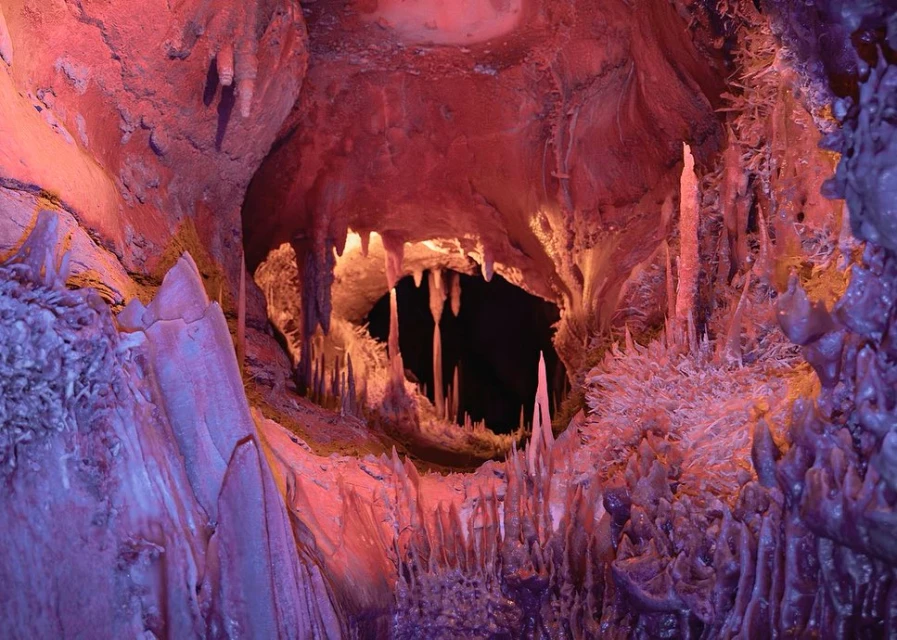 View of a cave with rock formations in hues of red and purple, illuminated by artificial light, creating a surreal landscape.