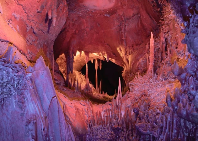 View of a cave with rock formations in hues of red and purple, illuminated by artificial light, creating a surreal landscape.