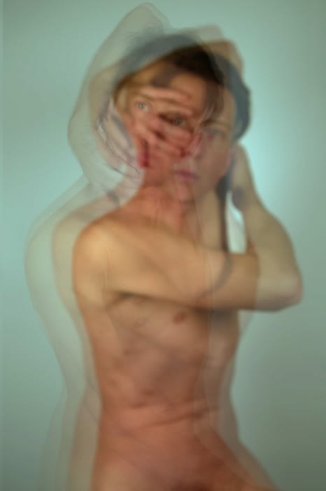 Abstract long exposure photograph of a nude adult with obscured face, against a pale blue background, creating a blurred, multi-layered effect.