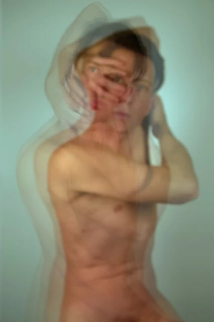 Abstract long exposure photograph of a nude adult with obscured face, against a pale blue background, creating a blurred, multi-layered effect.