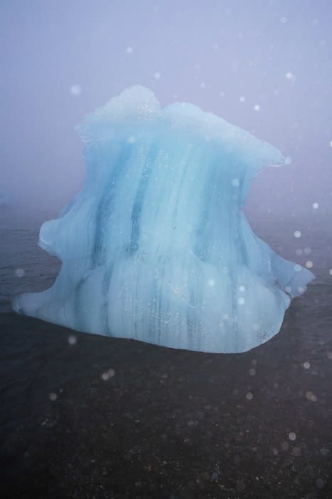 Icy blue iceberg floats on dark shoreline with snowy weather in landscape format, creating a serene and cold natural scene.