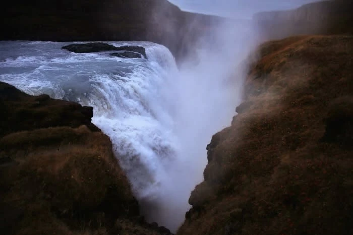 Waterfall cascading over rocks into a foggy gorge, surrounded by brown cliffs and vegetation. Natural scenic beauty in landscape format.